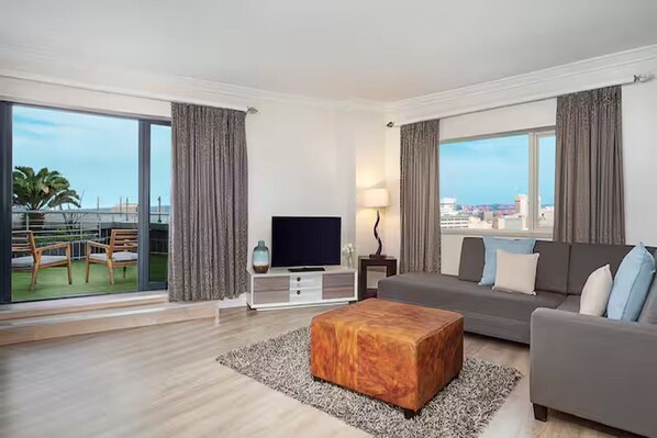 A modern hotel room with a large balcony, a flat-screen TV, a grey sofa, and a wooden ottoman. - Hilton Durban (Durban)