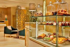 Un hall d’hôtel comprenant un espace salon, une vitrine où sont exposées des pâtisseries et des desserts, et un mur décoratif.