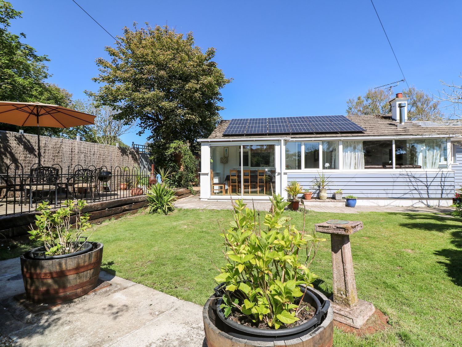 Een huis met een dakbedekt met zonnepanelen, een terras met stoelen en een tuin met planten in potten.