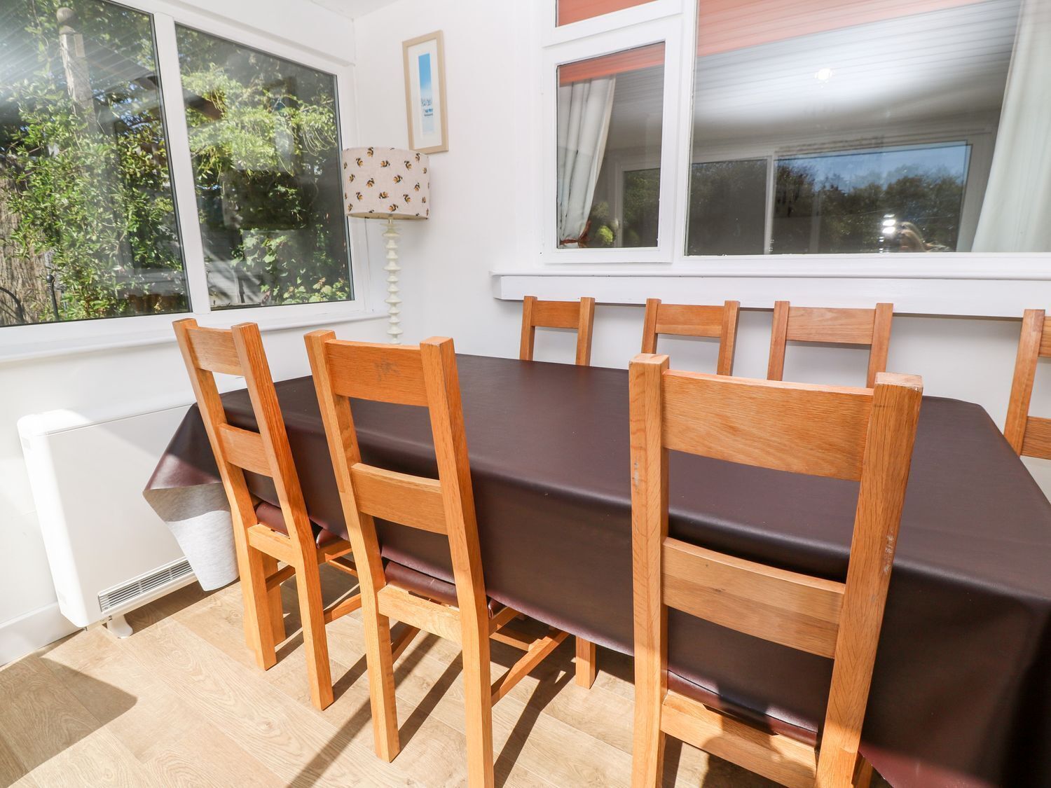 Uma sala de jantar com uma mesa retangular e seis cadeiras de madeira, janelas amplas com cortinas e um radiador.