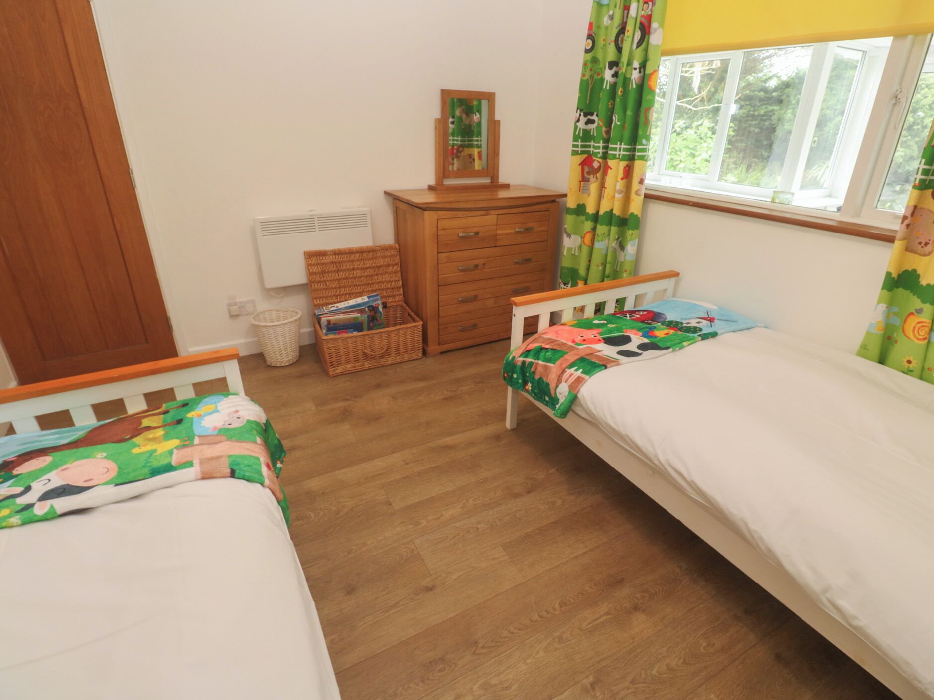 A bedroom with two single beds, a dresser, a wicker basket, and a window with curtains.