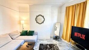 A modern living room with a sofa, a coffee table, a flat-screen TV displaying the Netflix logo, a floor lamp, and a mirror. - Spacious 4BR House + Garden Retreat | Parking | 7 people (Wallasey)