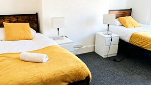Habitación de hotel con dos camas, cada una con cabecero de madera, ropa de cama blanca y almohada de color amarillo. Hay mesitas de noche con lámparas y un enchufe.