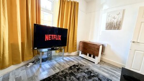 A living room with a television displaying the Netflix logo, a wooden cabinet, a patterned rug, and a window with curtains. - Spacious 4BR House + Garden Retreat | Parking | 7 people (Wallasey)