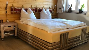 Ein Bett mit weißer Bettwäsche, ein Holz-Kopfteil, ein Nachttisch mit einer Uhr und ein Fenster mit einer Pflanze.