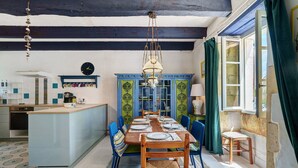 A dining area with a long table, chairs, a chandelier, and a kitchen in the background.