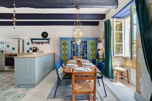 A dining area with a long table, chairs, a chandelier, and a kitchen in the background.