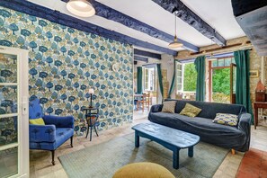 A room with a patterned wallpaper, a blue sofa, a wooden coffee table, and a dining area visible through an open door. - Holiday Home “Cottage Blanc” with Private Terrace and Wi-Fi (Beaumontois en Périgord)