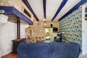 A rustic room with exposed beams, a stone wall, a bed with a blue cover, a wooden stove, and a patterned wallpaper. - Holiday Home “Cottage Blanc” with Private Terrace and Wi-Fi (Beaumontois en Périgord)
