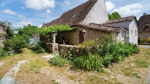 A rustic stone house with a sloped roof, a small garden with various plants, and a clear sky.