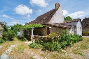 A rustic stone house with a sloped roof, a small garden with various plants, and a clear sky.