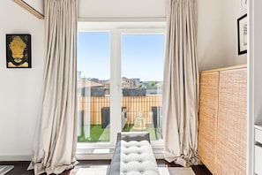 Ein Zimmer mit einer Schiebeglastür, die auf einen Balkon führt, einer gepolsterten Sitzbank, einem gewobenen Raumteiler und einem gerahmten Bild an der Wand.