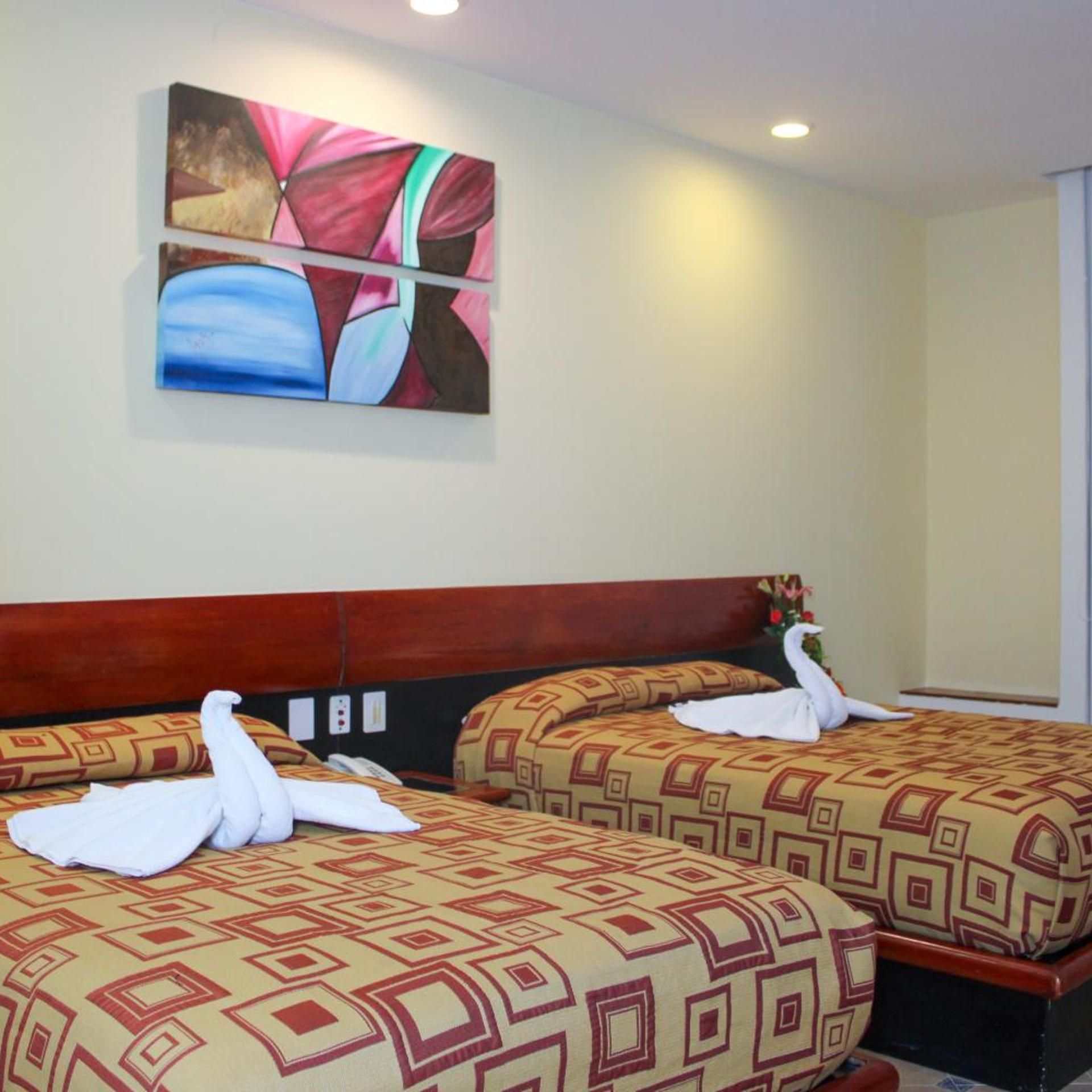 A hotel room with two beds, each with a patterned bedspread and a white towel shaped like a swan. There is a painting on the wall above the beds.