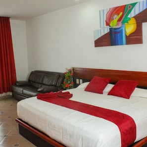 A hotel room with a bed, a red throw, a red pillow, a black leather sofa, a small plant, and a painting on the wall.