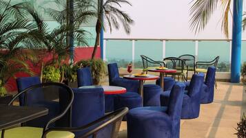 An outdoor seating area with round tables and blue cushioned chairs, surrounded by potted plants and overlooking a cityscape.