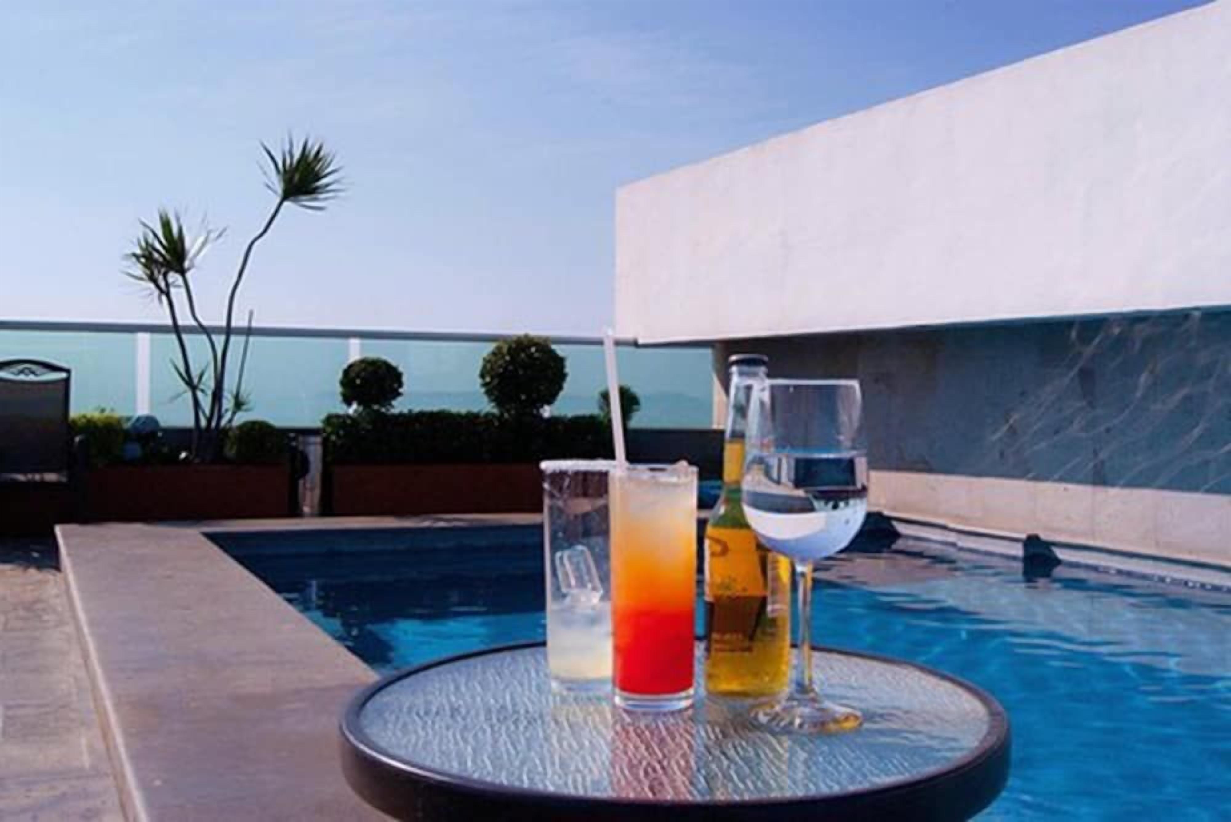 Escena junto a la piscina con una mesa redonda, un vaso de líquido rojo, un vaso de líquido blanco y una botella de líquido amarillo.
