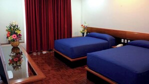 A hotel room with two beds, a wooden headboard, a red curtain, a small table with a vase of flowers, and a wooden floor.