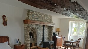A rustic room with a stone fireplace, a bed with patterned bedding, a wicker chair with cushions, and a dining area with wooden furniture. - Guest Room “Room 3” with Shared Pool, Communal Terrace and Wi-Fi (Saint-Bon)