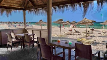 Un espace extérieur aménagé avec des tables et des chaises, donnant sur une plage parsemée de parasols et fréquentée par des gens.