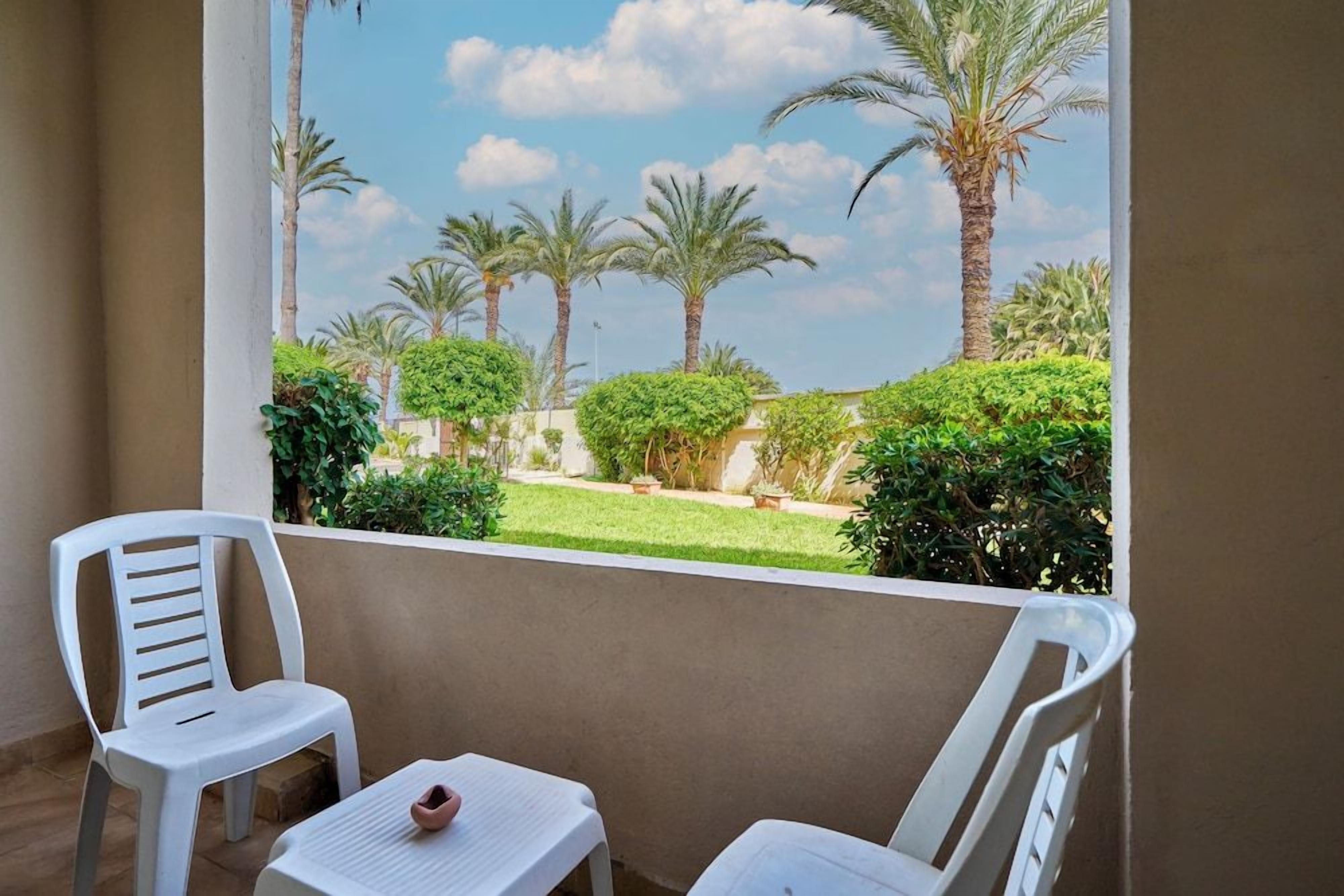 Un balcon avec deux chaises en plastique blanc et une petite table, donnant sur un jardin bien entretenu agrémenté de palmiers et d’arbustes.