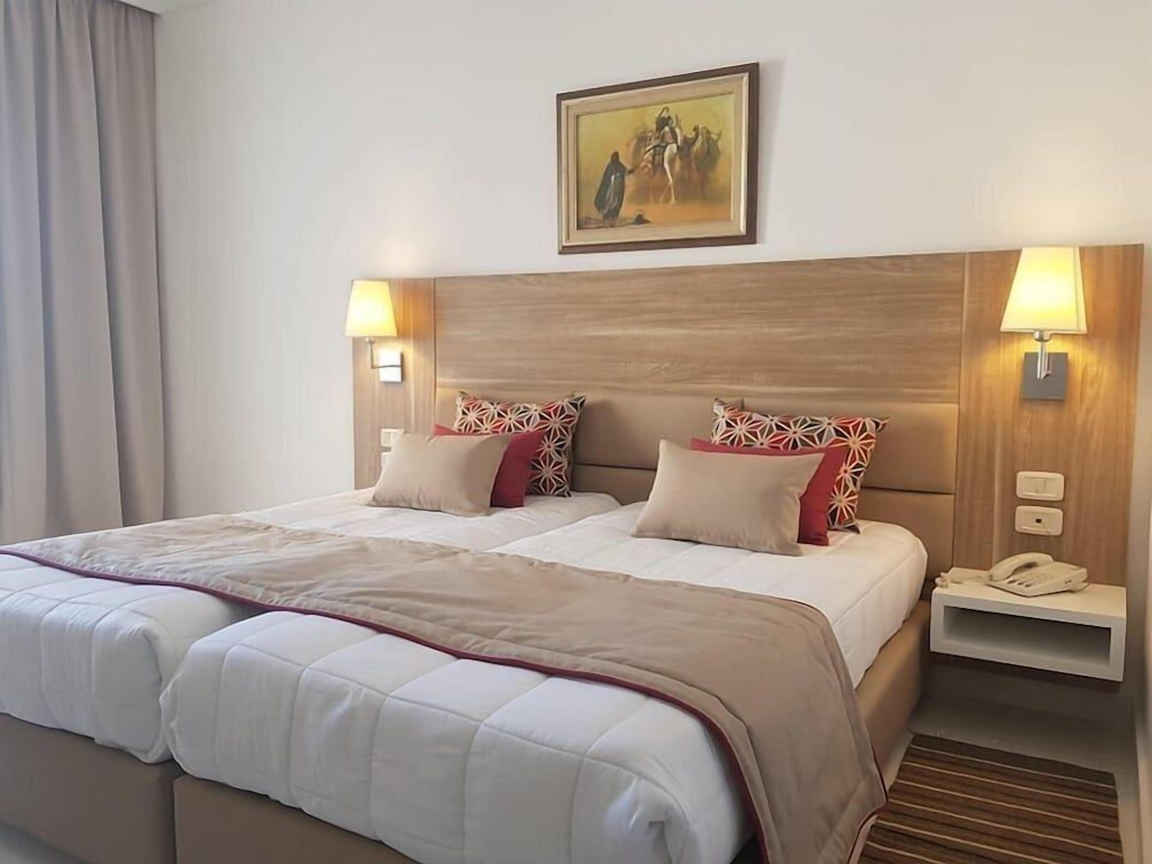A hotel room with a large bed, two bedside tables, a painting on the wall, and a lamp on each table.