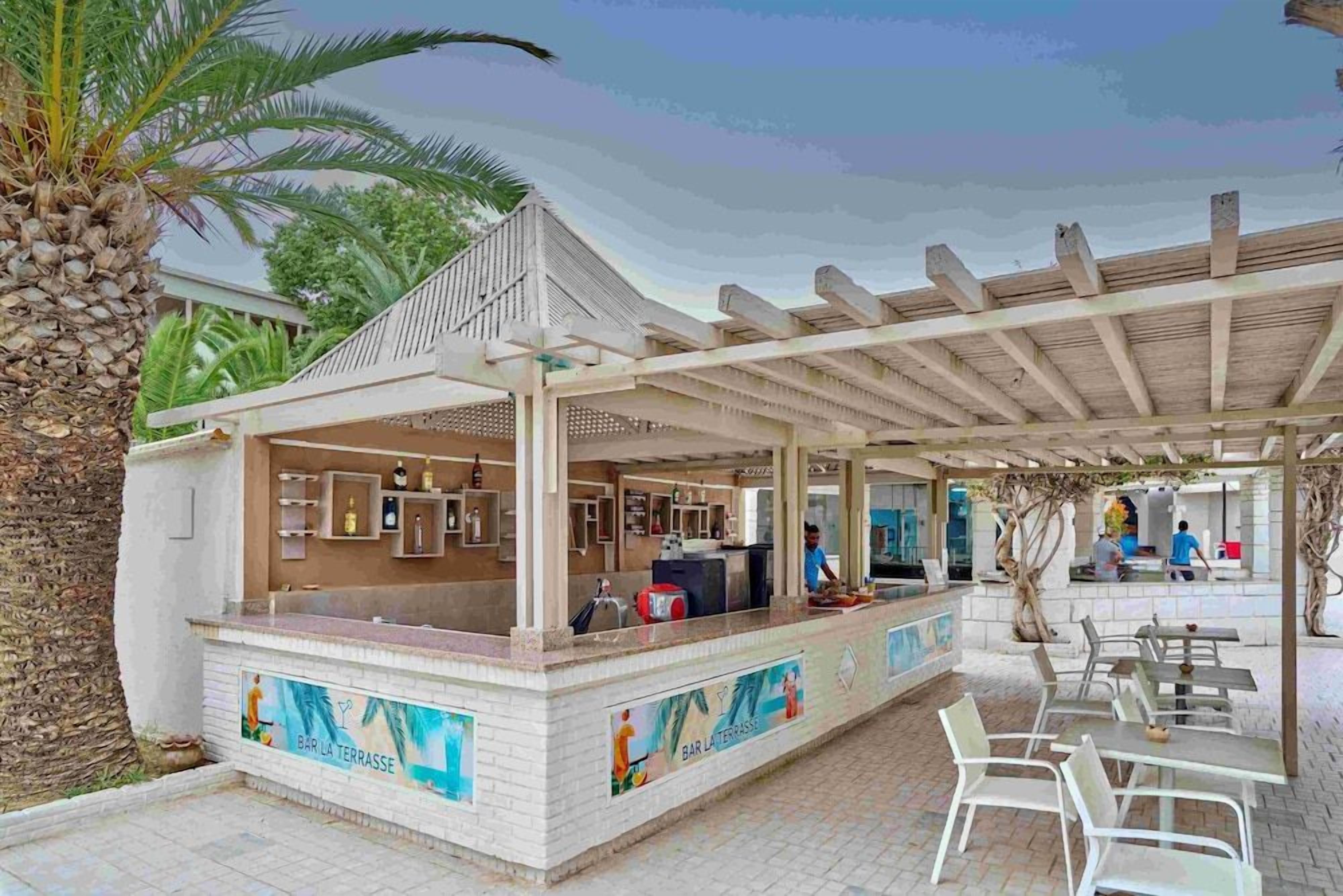 Uma área de bar ao ar livre com um espaço de estar coberto, balcão com cardápio e vista para a piscina.