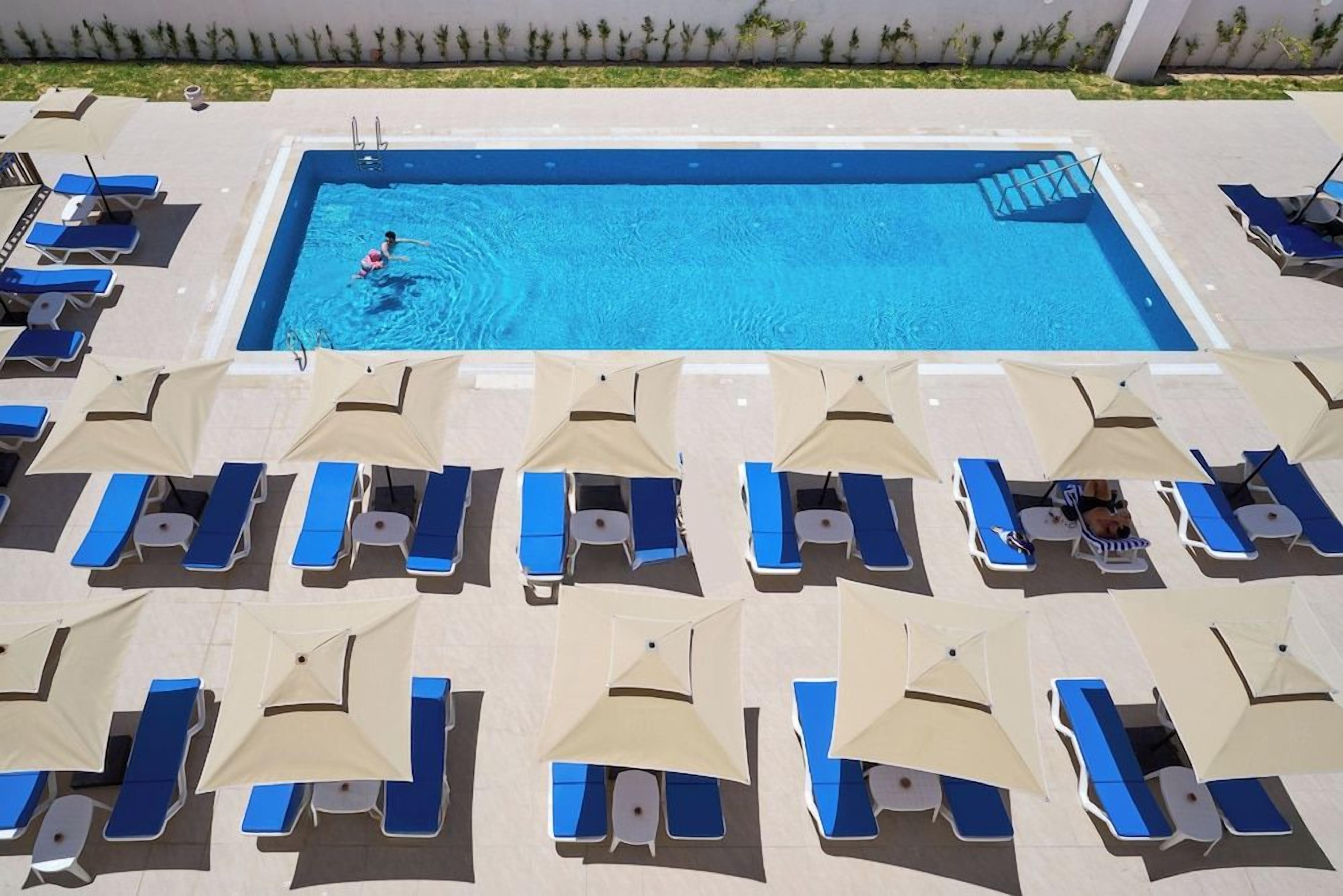 A rectangular swimming pool with a person swimming, surrounded by rows of blue lounge chairs and beige umbrellas.