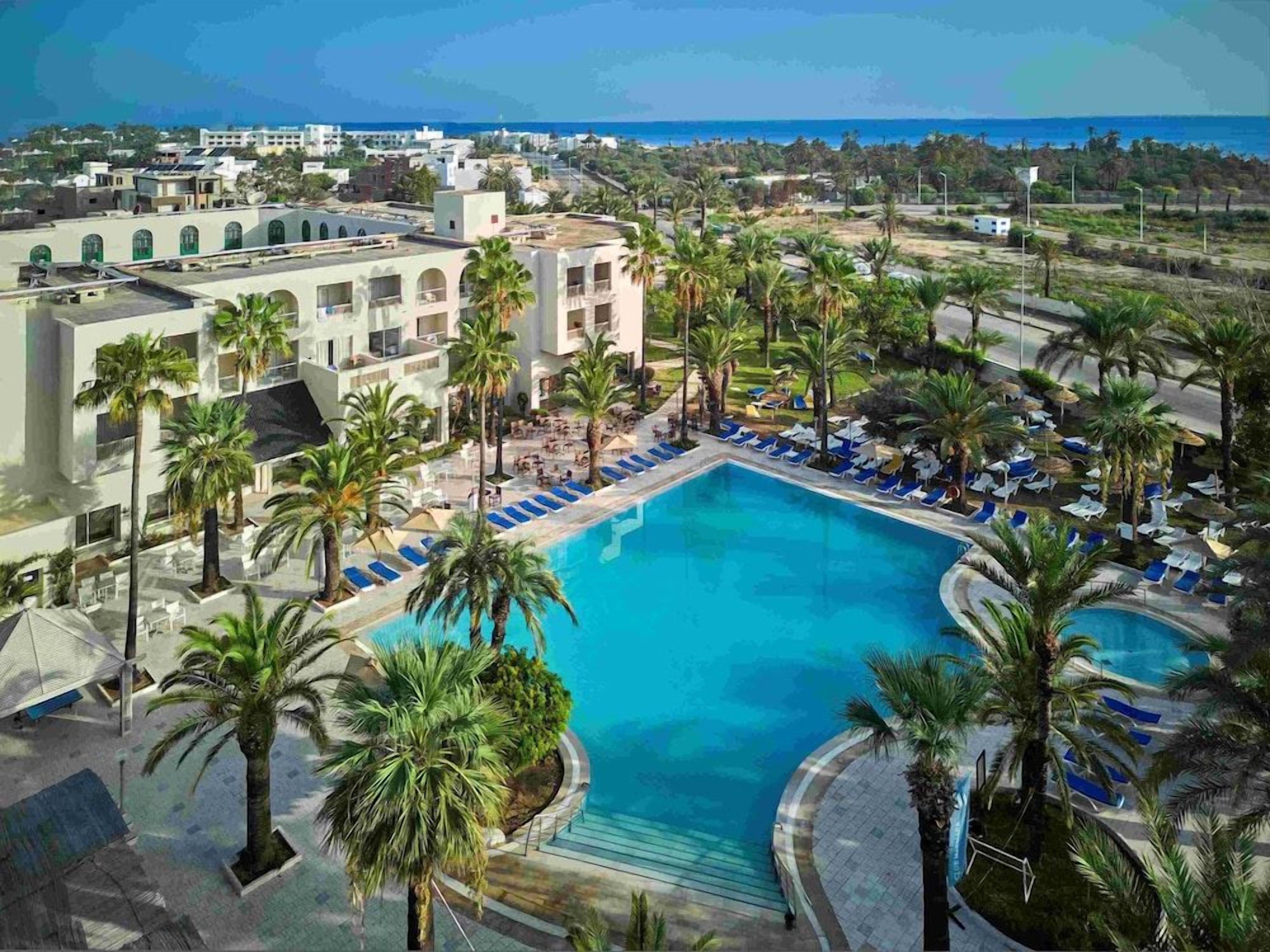 A resort with a central swimming pool surrounded by palm trees and lounge chairs.