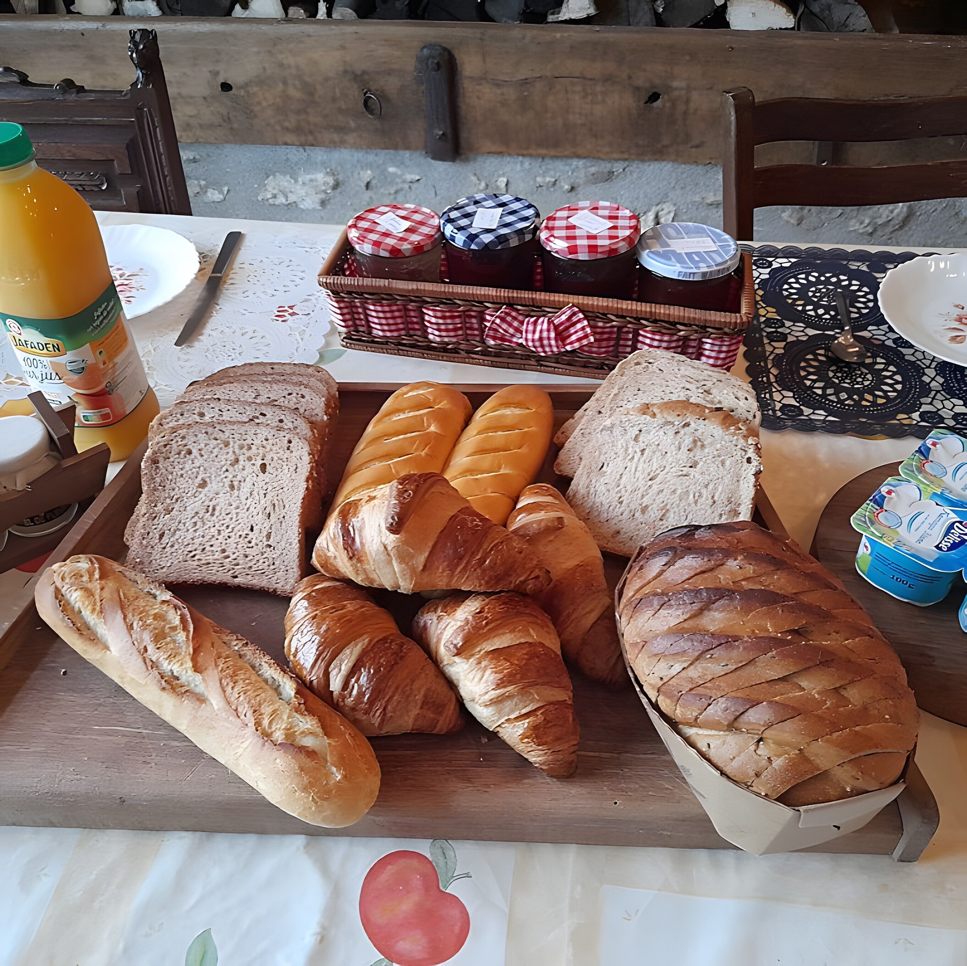 A variety of breads and jams on a wooden board, with a bottle of juice and a plate visible in the background.