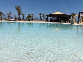 Piscina cristalina con suelo de baldosas, rodeada de palmeras y una estructura con sombra.
