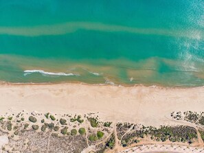 An aerial view of a beach with clear turquoise waters, sandy shore, and sparse vegetation.