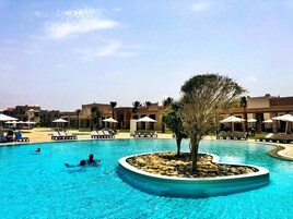 Una piscina con sillones de descanso y sombrillas, rodeada de edificios con estilo arquitectónico desértico.