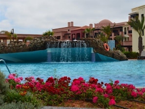 A swimming pool with a waterfall feature, surrounded by colorful flowers and a stone wall. - Oriental Bay Beach Golf and Spa Saida (Saidia)