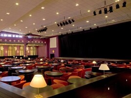 Un salón de conferencias con varias mesas y sillas, un escenario con cortina y una pantalla de proyector.