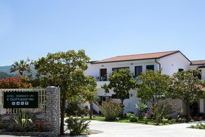 Il s’agit d’un hôtel à l’extérieur blanc, avec un toit recouvert de tuiles rouges et une enseigne indiquant qu’il s’agit d’une résidence. On y trouve des arbres et des plantes dans le jardin avant.