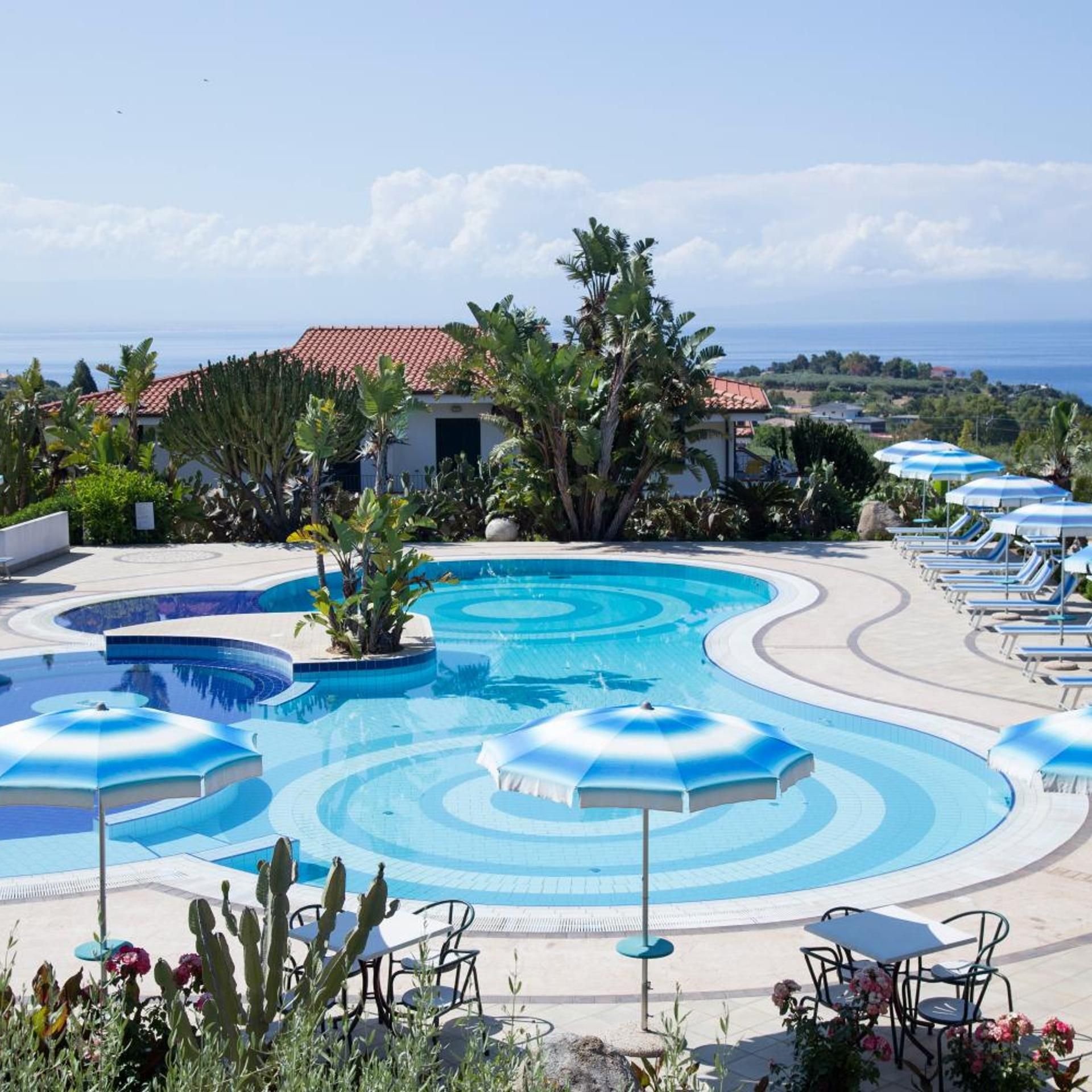 A pool with a wavy design, surrounded by lounge chairs and umbrellas, with a view of the sea and a building with a red roof in the background.