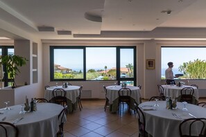 A dining area with round tables, chairs, and large windows offering a view of the sea. - Hotel Residence Il Gattopardo (Ricadi)
