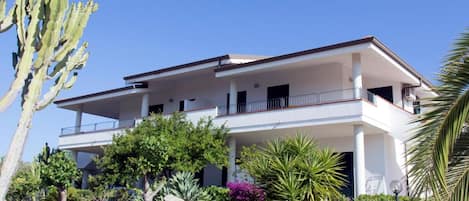 A two-story building with a balcony, surrounded by various plants and a cactus.