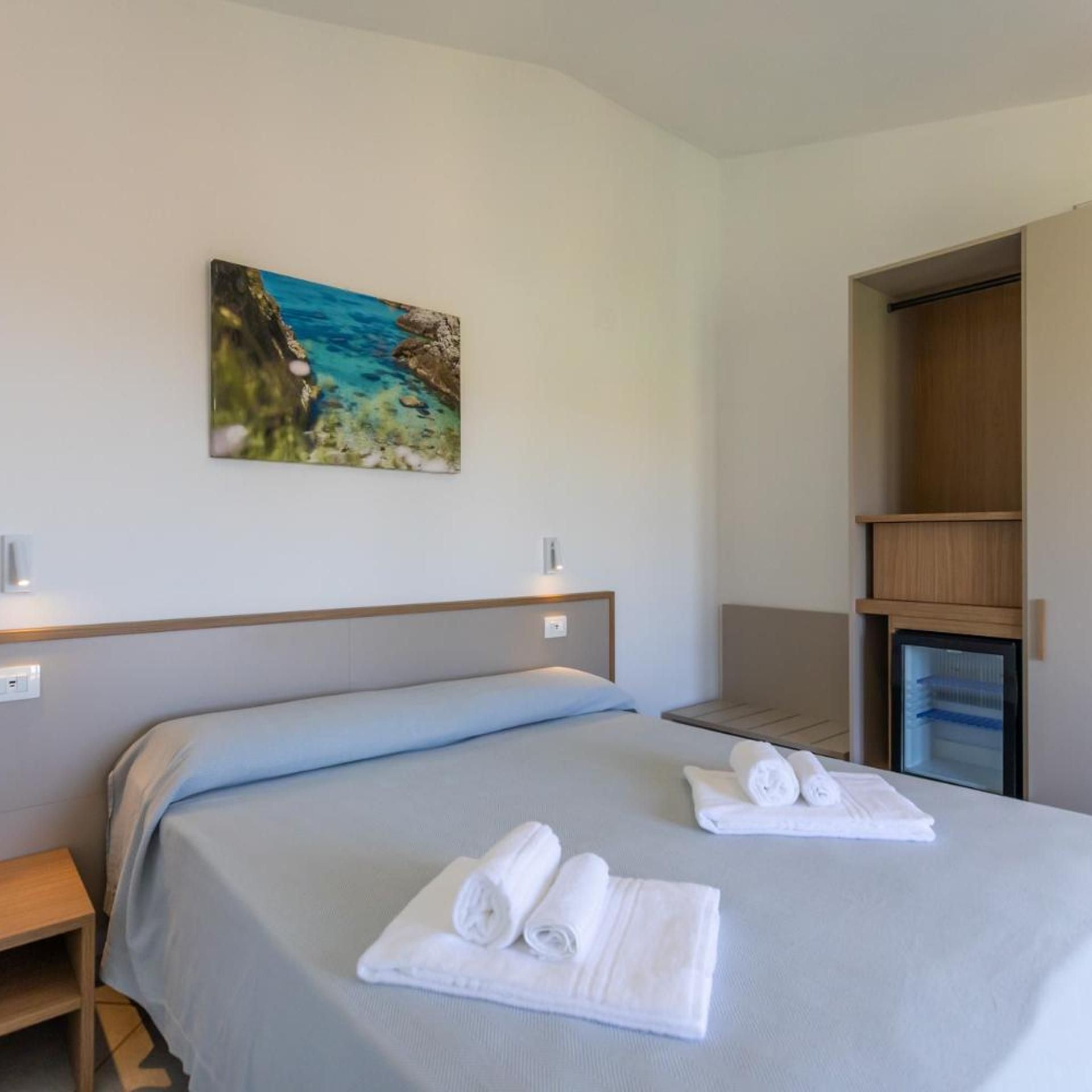 A hotel room with a bed, a small bedside table, a wall-mounted painting, a wardrobe, and a mini-fridge.