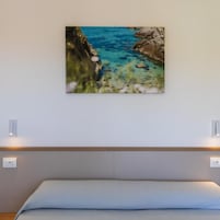 Un dormitorio con una cama, un cuadro colgado en la pared que muestra una escena costera y dos interruptores de luz instalados en la pared.