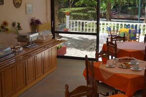 Un comedor para desayunos con barra libre, asientos al aire libre con manteles naranja y vistas a un jardín a través de un ventanal.