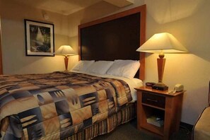 Un cuarto de hotel con una cama grande, dos lámparas de noche, una mesita de noche de madera con un teléfono y un cuadro en la pared.