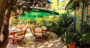 An outdoor seating area with wooden tables and chairs, green umbrellas, and potted plants. - Hotel Dalis (Nessebar)