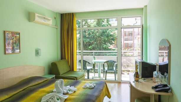 Hotelkamer met een bed, een bureau met een computer, een stoel, een tafel met stoelen en een raam met uitzicht op groen en gebouwen.