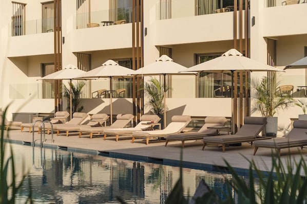 A poolside area with lounge chairs and umbrellas, a clear pool, and a building with balconies. - Inea Hotel and Suites (Chania)
