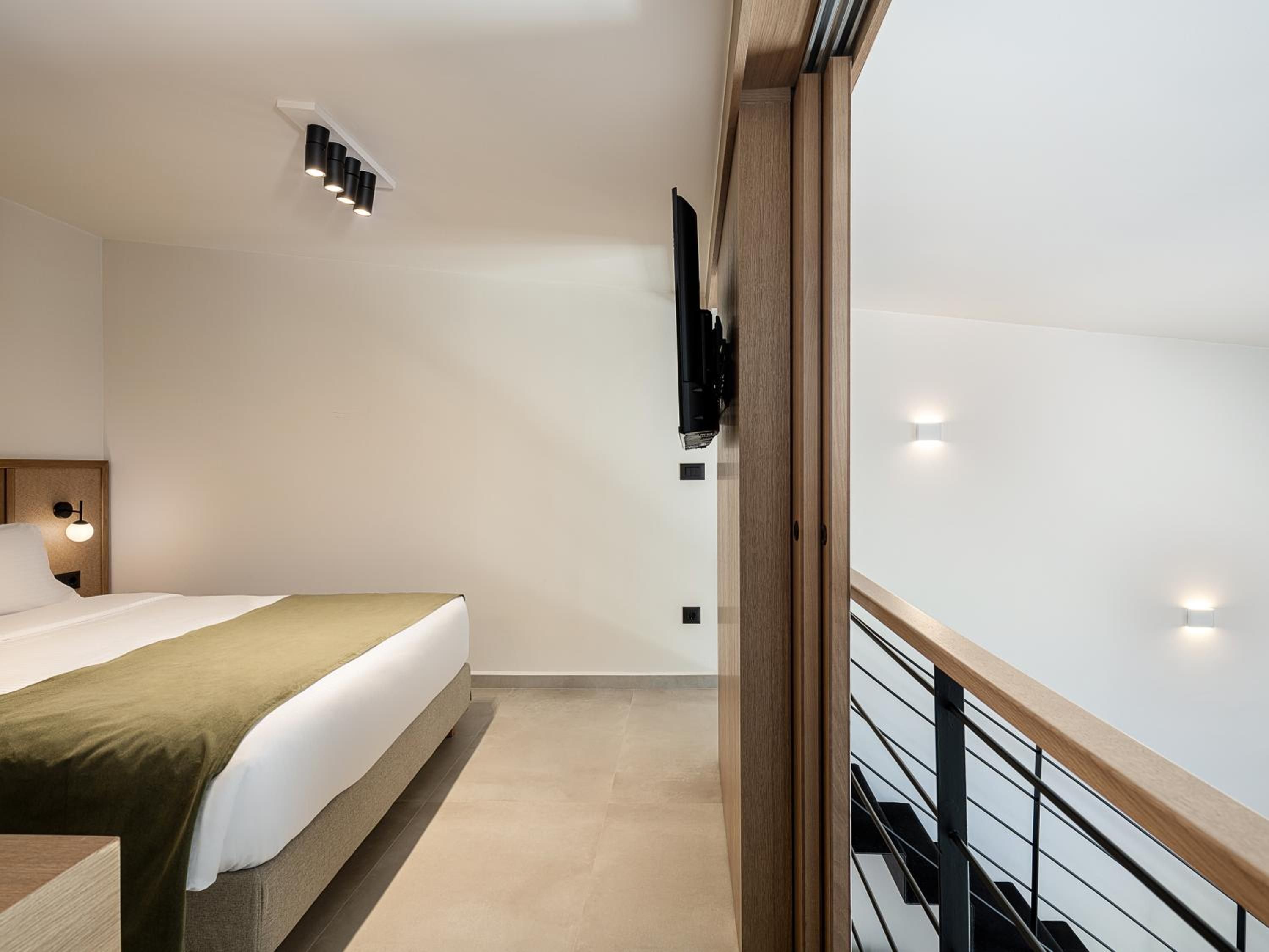 Une chambre d’hôtel moderne équipée d’un lit, d’une tête de lit en bois, d’un téléviseur à écran plat fixé au mur et d’un balcon doté d’une balustrade en bois.