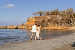 A couple walking on a sandy beach with a rocky cliff in the background. - Inea Hotel and Suites (Chania)