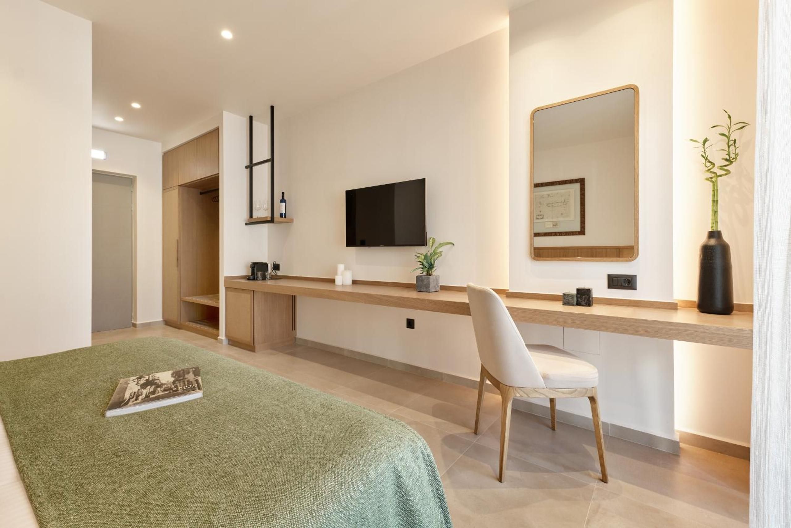 A modern hotel room with a bed, a desk, a chair, a flat-screen TV, a mirror, and a potted plant.