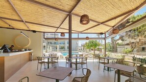 Un área de comedor al aire libre con mesas y sillas, una piscina al fondo y vistas a edificios y zonas verdes.