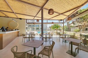 Un área de comedor al aire libre con mesas y sillas, una piscina al fondo y vistas a edificios y zonas verdes.